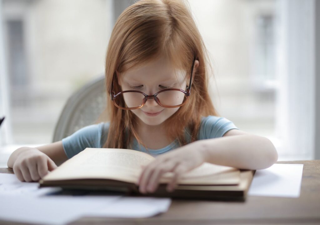 A focused young girl in glasses reads a book indoors, reflecting education and concentration.